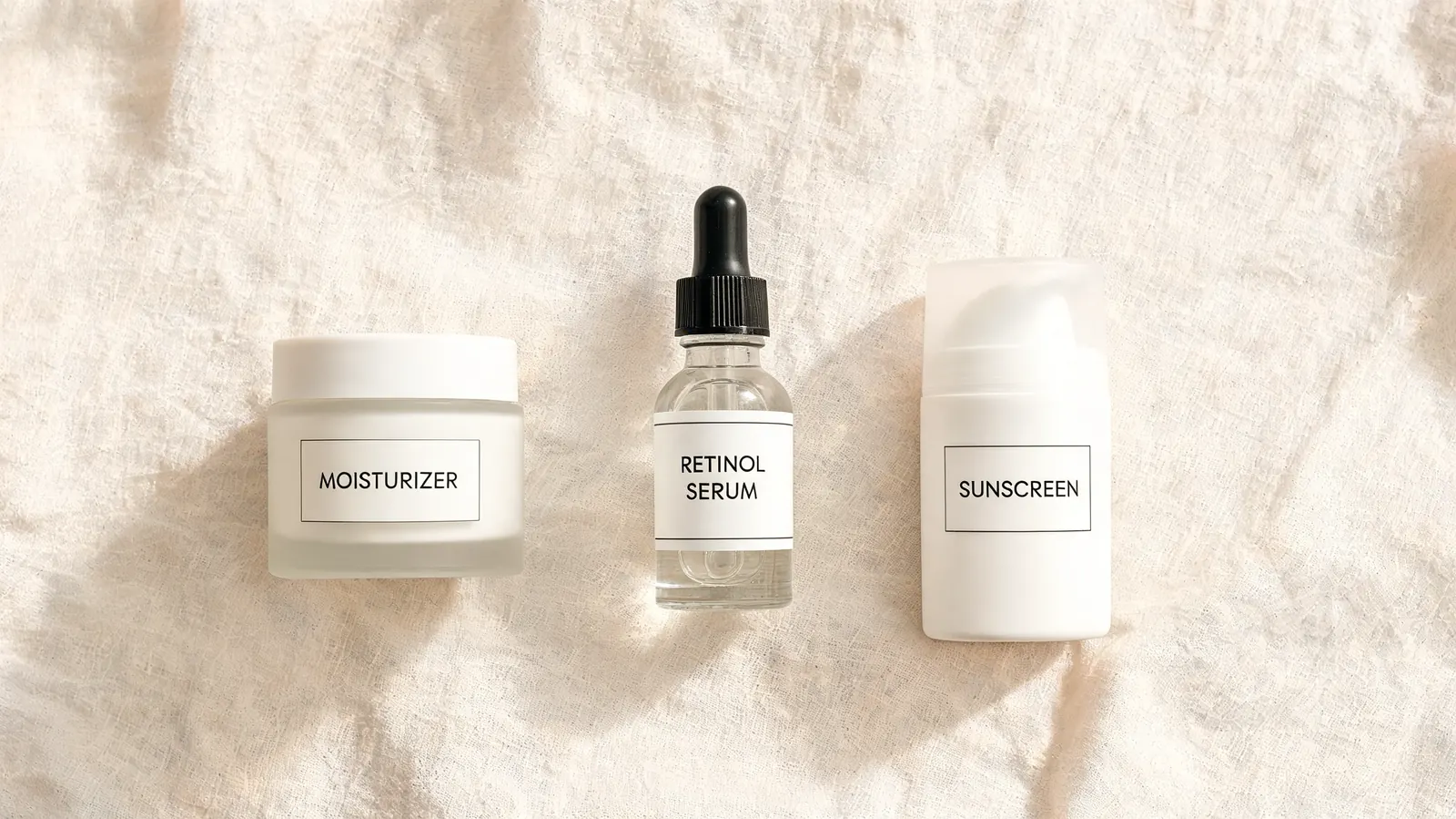 Overhead flat lay of retinol serum bottle, a small moisturizer tube, and a plain white sunscreen bottle on a cream linen surface