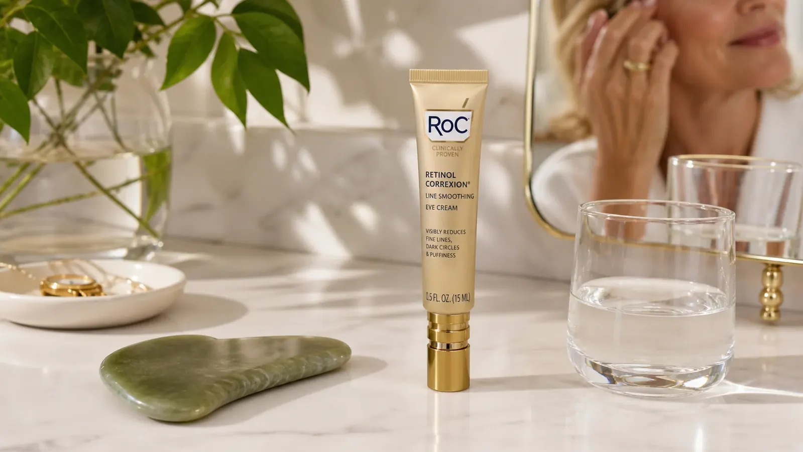 Close-up of a small tube of RoC Retinol Correxion Under Eye Cream resting on a marble bathroom counter beside a jade gua sha tool and a glass of water