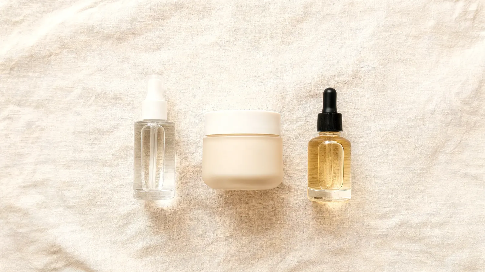Flatlay of a simple morning skincare routine on a cream linen surface: vitamin C serum, moisturizer, and SPF side by side